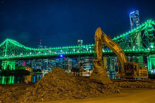 Excavator heavy equipment on shore of glowing modern city digging soil in night time, Brisbane